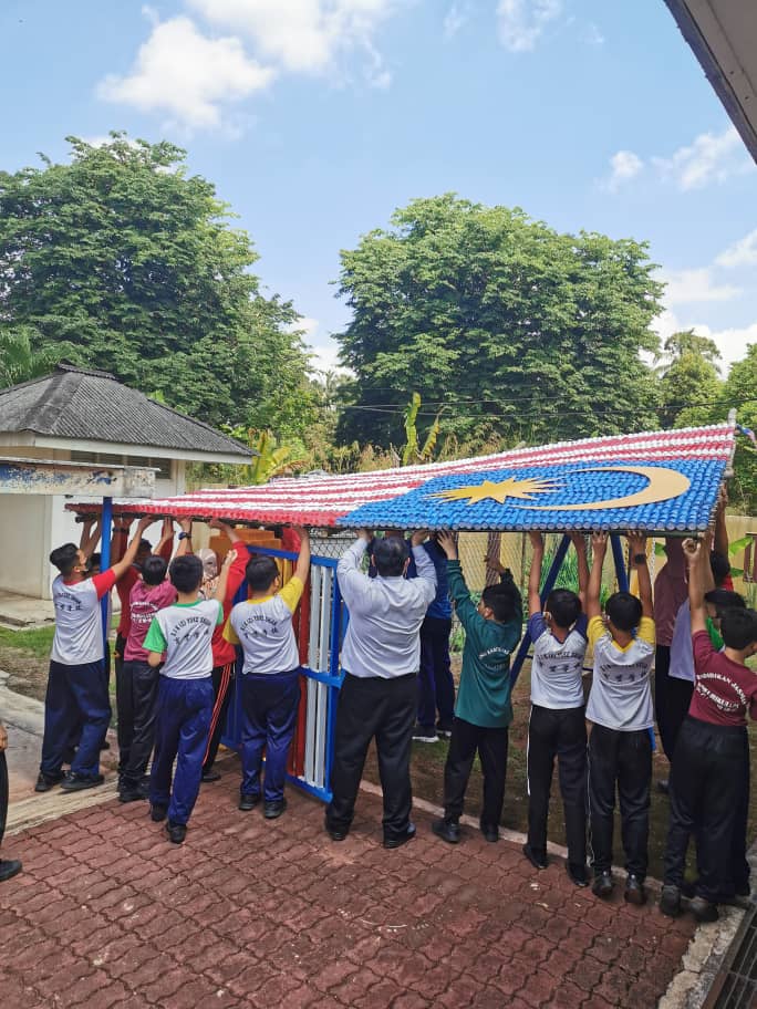 关丹育贤华小约300名师生，使用近千个塑料益生菌瓶子，费时两周，制作出10公尺乘14公尺的巨型国旗，让学生感受绿色国庆日，别具意义。